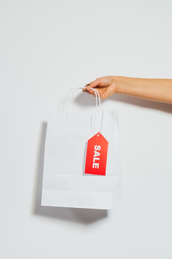 A persons hand holding a white shopping bag with a red sale tag on a white background.